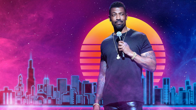 Deon Cole on stage, mid-performance, arms outstretched as if inviting the audience into a secret