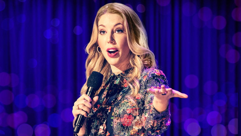 Katherine Ryan on stage, bathed in soft pink and purple lighting, gesturing with a microphone in hand