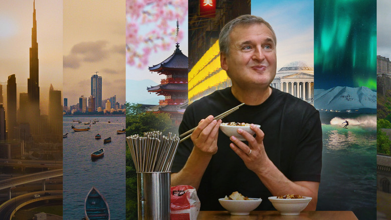 Phil Rosenthal sitting at a table in a bustling market, surrounded by plates of food, grinning at the camera