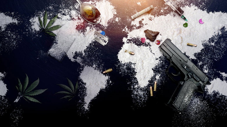A masked figure in shadow, their face obscured by a balaclava, stands in a dimly lit room filled with bags of drugs.