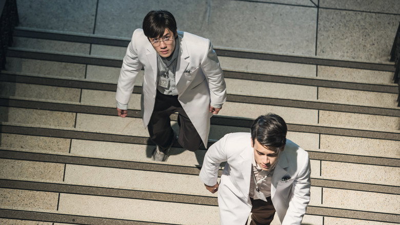 Two doctors stand in a dimly lit hospital corridor, their faces half-lit by the glow of medical equipment, tension etched into their expressions.