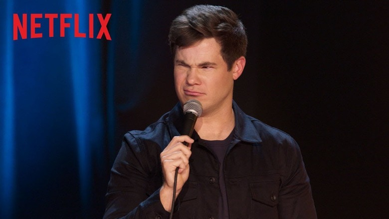 Adam Devine on stage, mid-performance, arms outstretched as he engages with the audience