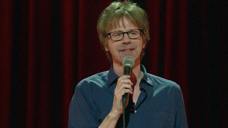 Dana Carvey on stage, mid-performance, bathed in warm light, a microphone in hand