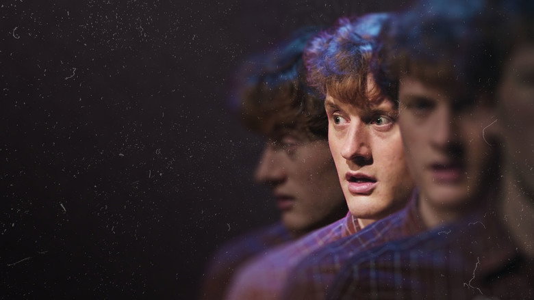 A dimly lit stage with a single spotlight on James Acaster, mid-gesture, as he delivers a punchline with exaggerated earnestness.