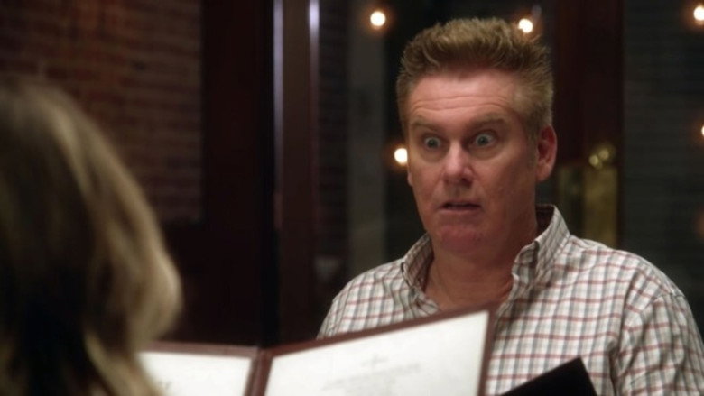Brian Regan mid-performance, gesturing with his hands as he delivers a joke, the audience blurred in the background