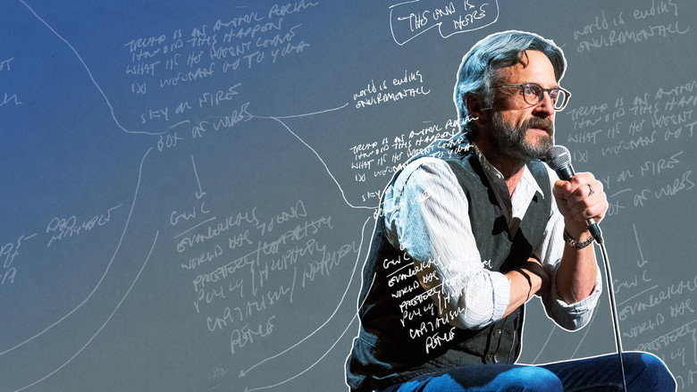 Marc Maron on stage, bathed in dim light, mid-monologue, his expression a mix of exhaustion and wry amusement.