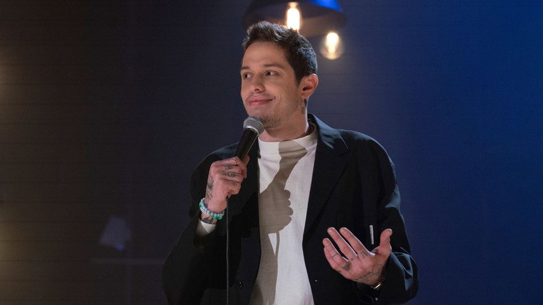 Pete Davidson on stage, mid-gesture, under warm stage lights