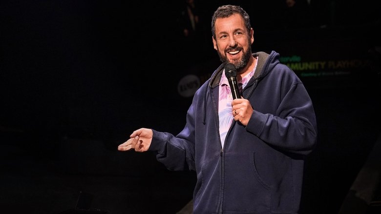 Adam Sandler on stage in a dimly lit, rundown theater, microphone in hand, looking slightly disheveled but grinning.