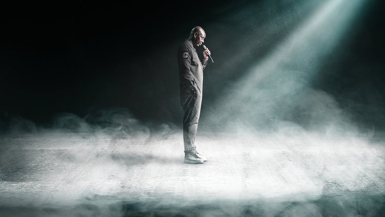 Chappelle on stage, mid-performance, his expression unreadable under the spotlight