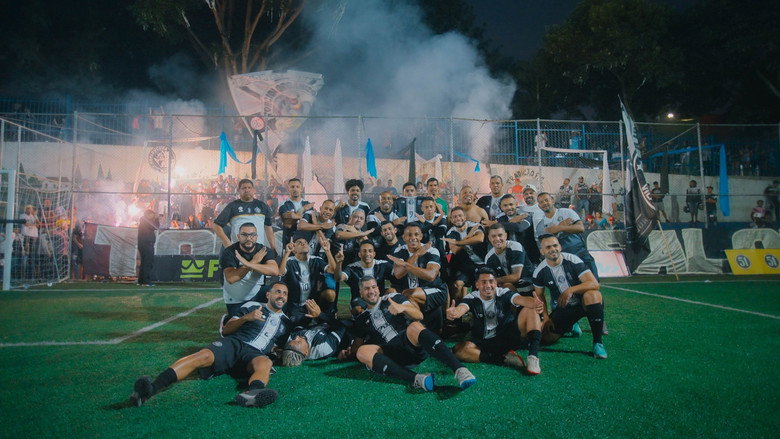 A drone shot of a dirt pitch in São Paulo, surrounded by favelas, with players mid-game under a hazy sky.