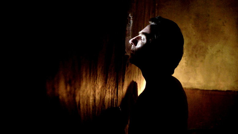 A dimly lit room, the only light seeping through a crack in the wall, casting long shadows across Higinio’s face as he presses his ear to the plaster.