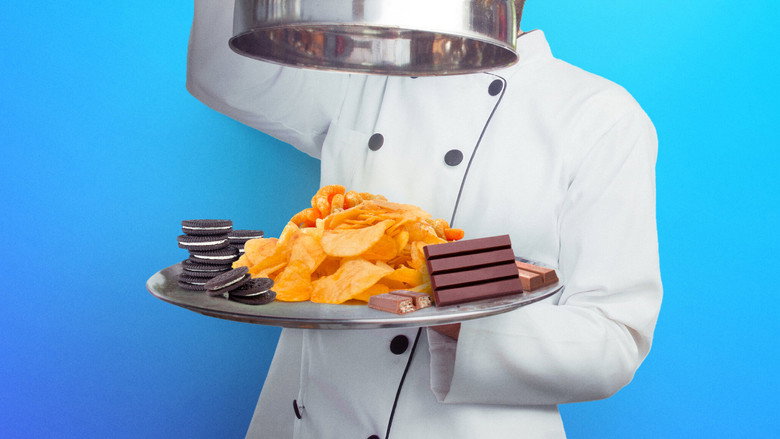 A chef in a bright kitchen holds up a plate of meticulously reconstructed snack food, the tension between homemade craft and mass-produced familiarity written in their focused expression.