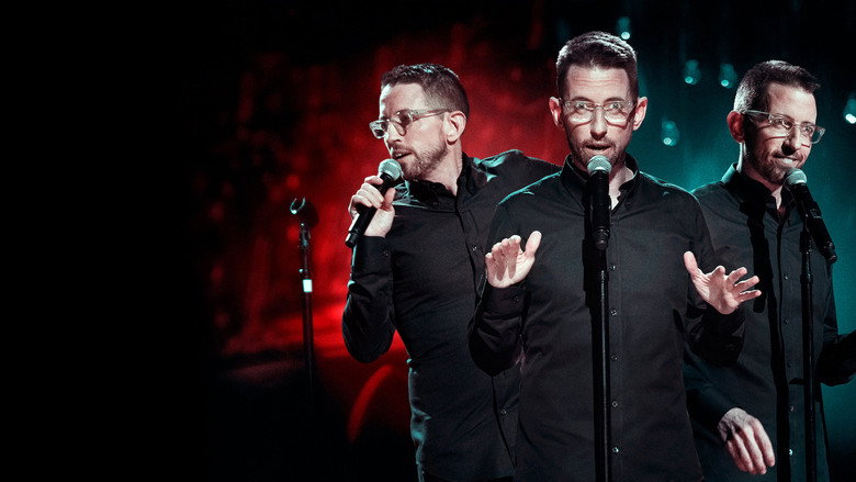 Neal Brennan on stage, bathed in blue light, holding a microphone like it’s the only thing keeping him upright