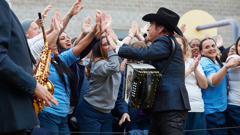 Los Tigres del Norte performing on stage at Folsom Prison, the band in black suits against a backdrop of inmates in khaki uniforms