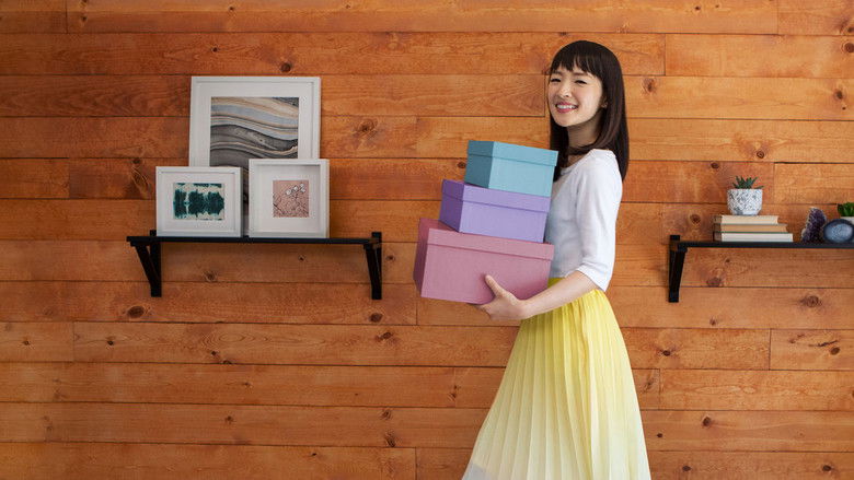 Marie Kondo kneels on the floor of a cluttered living room, hands pressed together in a moment of quiet reflection before beginning the tidying process.