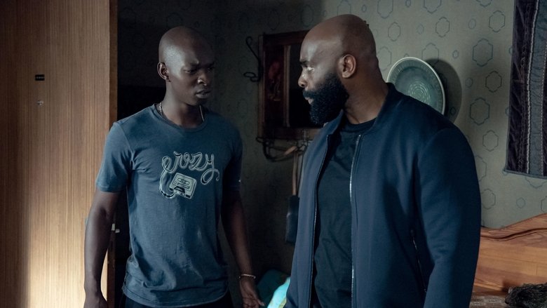 Two brothers stand in a dimly lit room, one facing the camera with a hardened expression, the other turned away, his posture tense and uncertain.