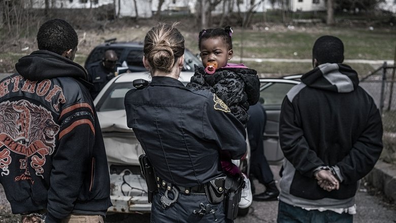 A Flint police officer sits in his cruiser at night, the dashboard lights casting a blue glow over his weary face.