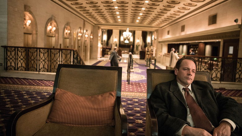 A man in a suit stands in a dimly lit room, his face half in shadow, staring at something unseen.