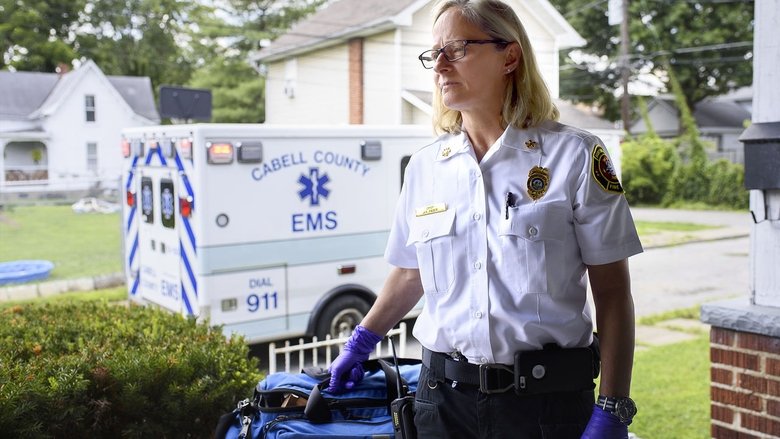 A fire chief kneels beside an overdose victim in the back of an ambulance, her gloved hands pressing an oxygen mask to their face.