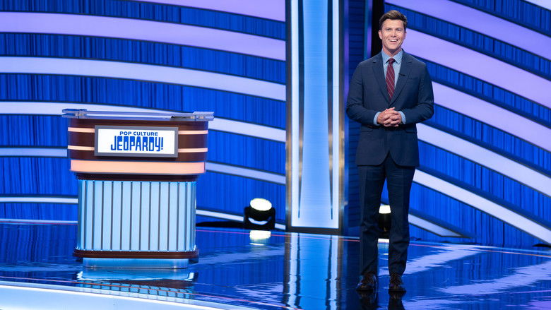 The LED-heavy set of *Pop Culture Jeopardy!*, bathed in neon blues and purples, with Colin Jost standing at the podium, microphone in hand, looking equal parts amused and exhausted.