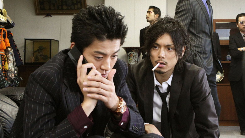 Two detectives stand in a dimly lit alley, their breath visible in the cold air, one leaning against a wall with his arms crossed, the other staring intently at something off-camera.