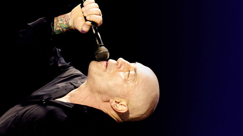Joe Rogan mid-performance, gesturing with his hands, the audience blurred in the background