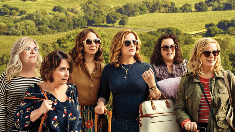 A group of women sit around a table in a sunlit vineyard, glasses of wine in hand, laughing as one of them gestures wildly.