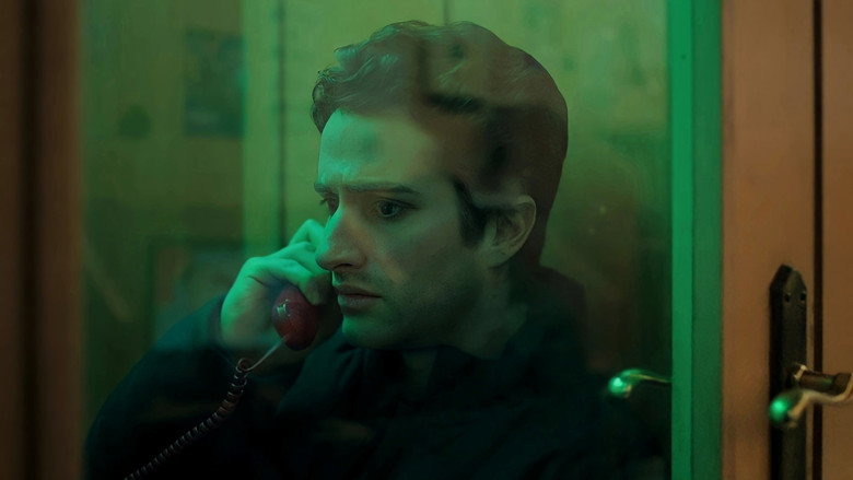 A dimly lit room where Juan Pablo (Dario Yazbek Bernal) sits at a desk, his face half in shadow, staring at a laptop screen with an expression caught between exhaustion and manic energy.