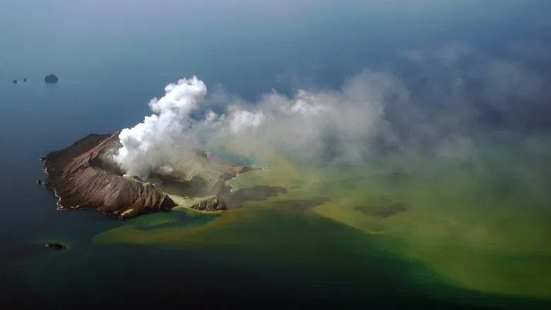 A helicopter hovers over the smoldering crater of Whakaari, its rotors kicking up plumes of gray ash against a stark blue sky.