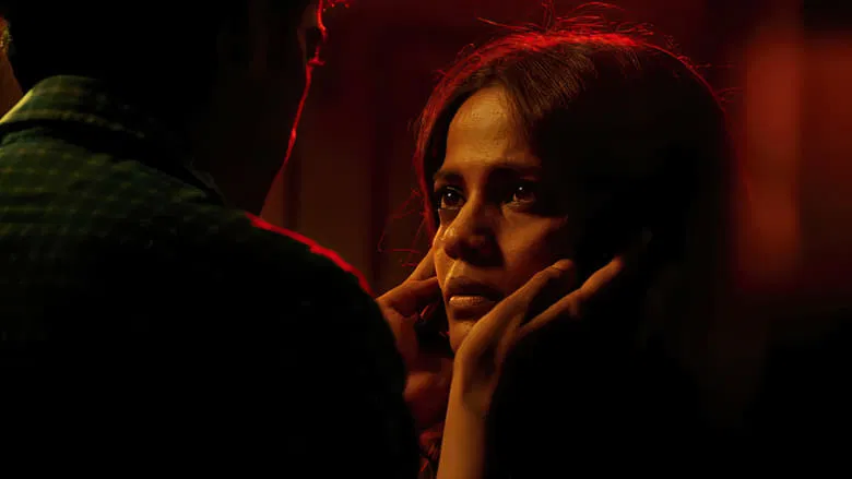 A dimly lit room where a woman in a sari sits on a bed, her face half in shadow, staring at something—or someone—off-camera.