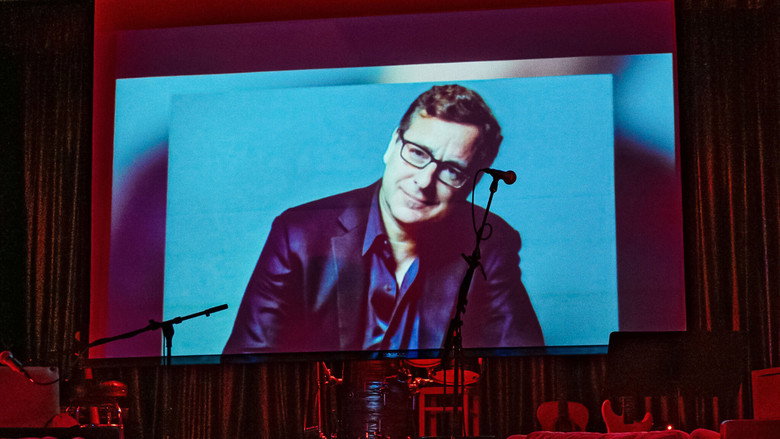 A dimly lit stage at the Comedy Store, the microphone stand glowing under warm lights, the audience blurred in the foreground.