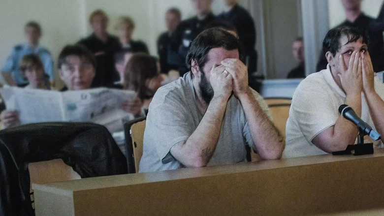 A dimly lit courtroom, the camera focused on the backs of the accused, their faces obscured by shadow