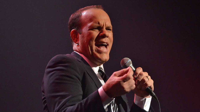 Tom Papa on stage, mid-laugh, bathed in warm light, the audience a blur of faces in the background.