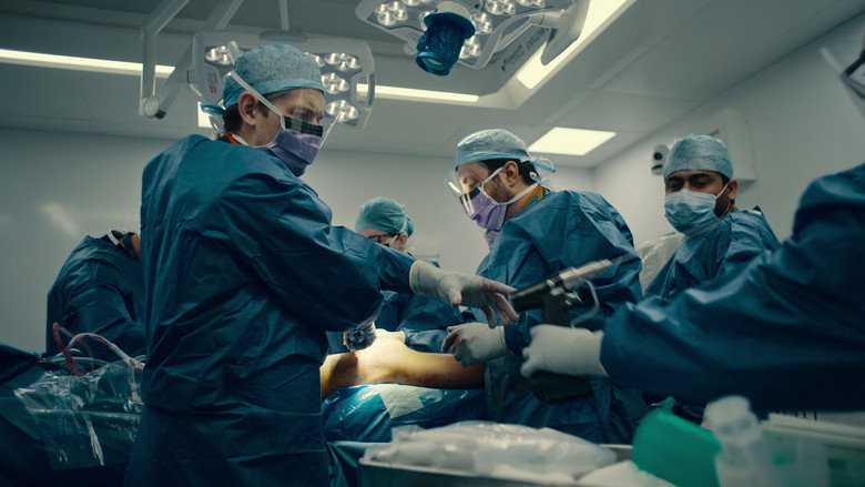 A trauma team in blue scrubs and gowns working urgently over a patient on a gurney, their faces tense under the fluorescent lights.