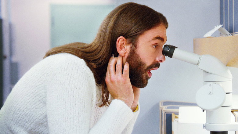 Jonathan Van Ness mid-conversation, gesturing with his hands, a bookshelf and warm lighting in the background