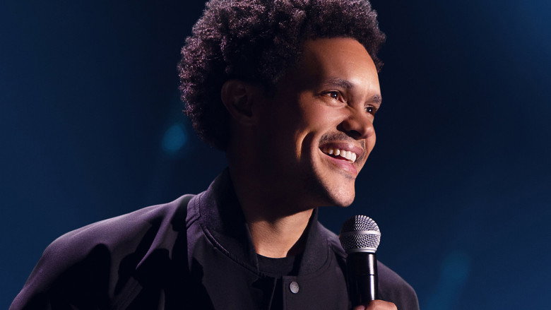 Trevor Noah on stage, mid-gesture, bathed in warm light, the audience blurred in the background