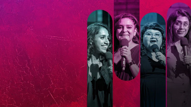 Four women stand on a dimly lit stage, microphones in hand, bathed in warm spotlight. The audience is a blur of faces, leaning in but not quite laughing.