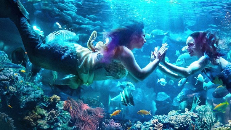 A mermaid performer in a shimmering tail, suspended underwater in a tank, her arms outstretched as if embracing the weight of the water.