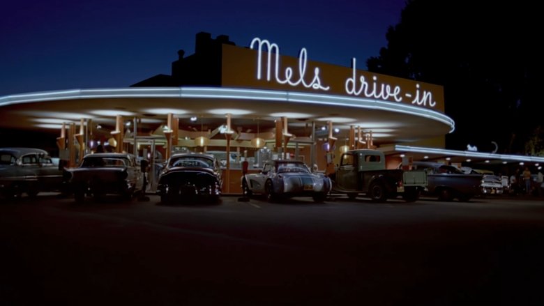 A neon-lit street at night, cars lined up along the curb, headlights cutting through the warm California haze.