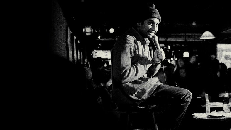 Aziz Ansari onstage, mid-gesture, under warm stage lighting