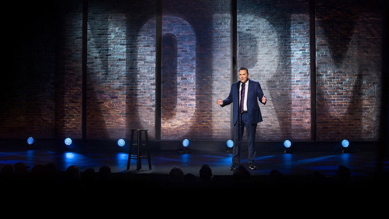 Norm Macdonald on stage, mid-performance, his expression deadpan as he delivers a joke.