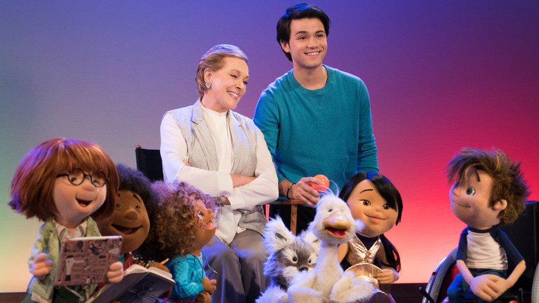 Julie Andrews stands in a bright, colorful classroom, surrounded by a group of expressive puppet students, all looking up at her with eager faces.