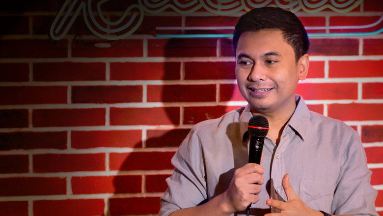 A dimly lit comedy club stage, where Raditya Dika’s character, Panca, stands under a single spotlight, his posture slumped as he delivers a joke to a nearly empty room.