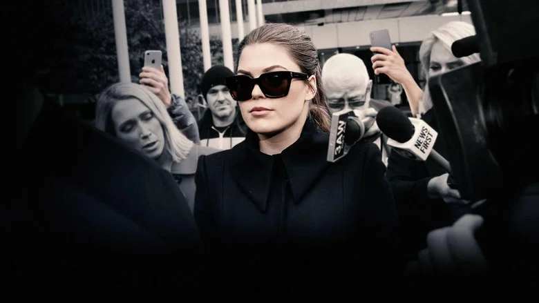 A close-up of Belle Gibson’s face, half in shadow, her expression unreadable as she speaks to an unseen interviewer.