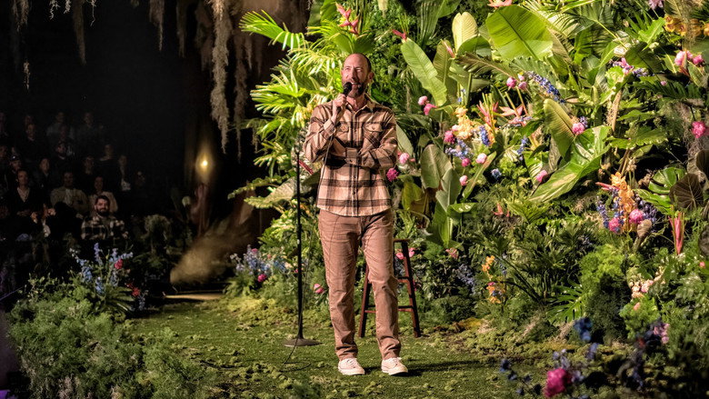 Ari Shaffir on stage, mid-gesture, with a backdrop of lush greenery that contrasts sharply with his dark suit and the dim lighting.