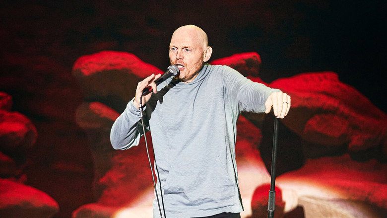 Bill Burr on stage at Red Rocks, bathed in warm light, the vast amphitheater stretching behind him