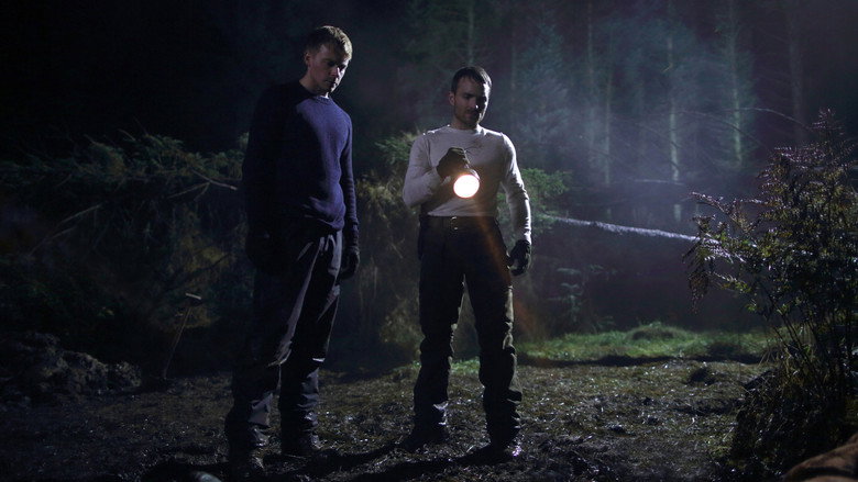 Two men stand in a dimly lit room, one holding a rifle, the other looking away with a tense expression