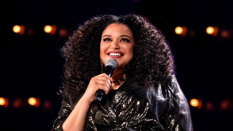 Michelle Buteau on stage, mid-laugh, gesturing with her hands as she delivers a punchline