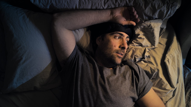 A dimly lit room where two characters sit on opposite ends of a couch, their faces half in shadow, the space between them charged with unspoken tension.
