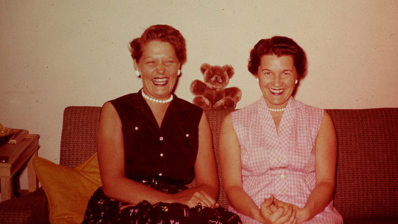 Two elderly women sit side by side on a couch, looking at old photographs in their laps.
